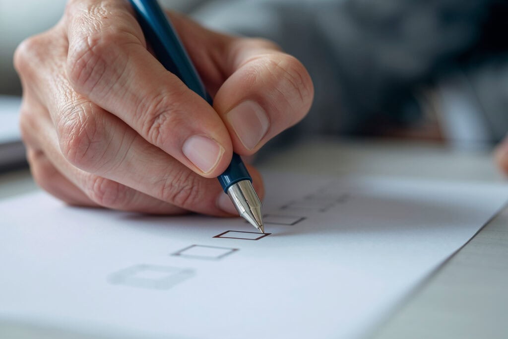 An elderly hand checks off boxes on a checklist with blue ball point pen before buying a mattress topper.